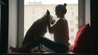 Genç bir kadın pencere eşiğinde oturmuş köpeğiyle konuşuyor ve oynuyor.