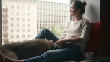 Genç bir kadın evinin pencere eşiğinde oturmuş köpeğini okşuyor.