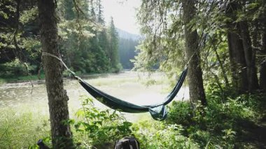 Boş bir hamağın sinematik çekimi bir yaz günü bir dağ gölünün arka planında rüzgarda sallanıyor.