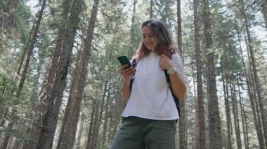 Sırt çantalı genç bir kadın yaz ormanında yürüyüş yaparken akıllı telefonunu kullanıyor.