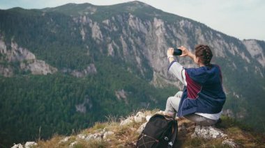 Dişi bir gezgin dağın tepesinden çarpıcı bir manzarayla otururken akıllı telefonunu kullanır.