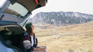 A young woman uses a smartphone while sitting in the trunk of her car with a stunning view of the mountains