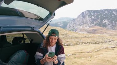 A young woman uses a smartphone while sitting in the trunk of her car with a stunning view of the mountains