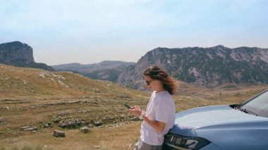 A beautiful young woman uses a smartphone while traveling by car through the mountains