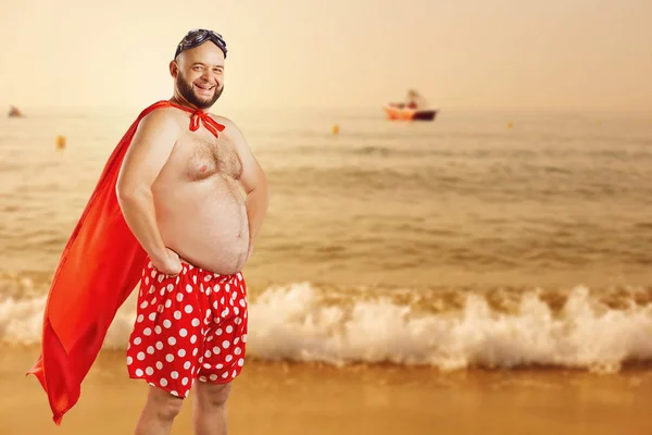 Engraçado homem gordo em um traje de super-herói na praia no verão — Fotografia de Stock
