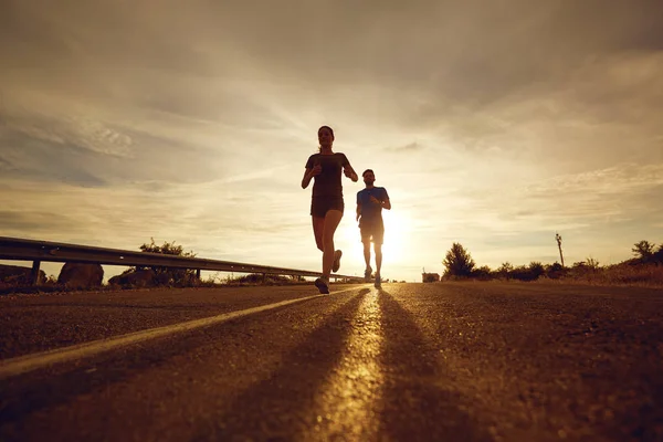 Ein Mann und ein Mädchen joggen bei Sonnenuntergang in der Natur entlang der Straße. — Stockfoto