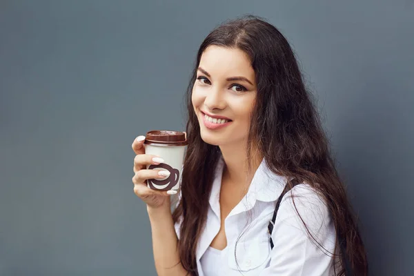 Ein brünettes Mädchen mit einem Kaffeebecher vor grauem Hintergrund. — Stockfoto