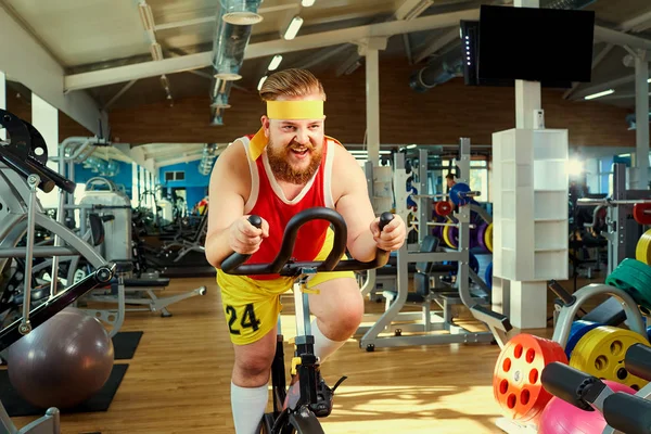 A funny fat man  doing exercises on an exercise bike in a sports