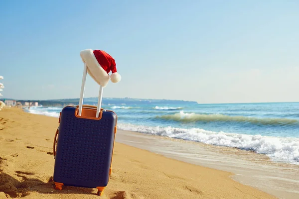 Sombrero de Santa Claus en una maleta en la playa junto al mar. Navidad —  Fotos de Stock