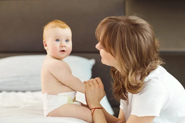 Gelukkig moeder met baby zittend op het bed binnenshuis. — Stockfoto