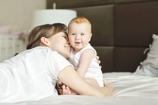 Gelukkig moeder knuffelen baby liggend op het bed binnenshuis. — Stockfoto