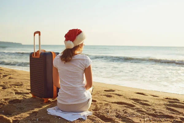 A girl in a Santa Claus hat with a suitcase on the beach on Chri — Stock Photo, Image