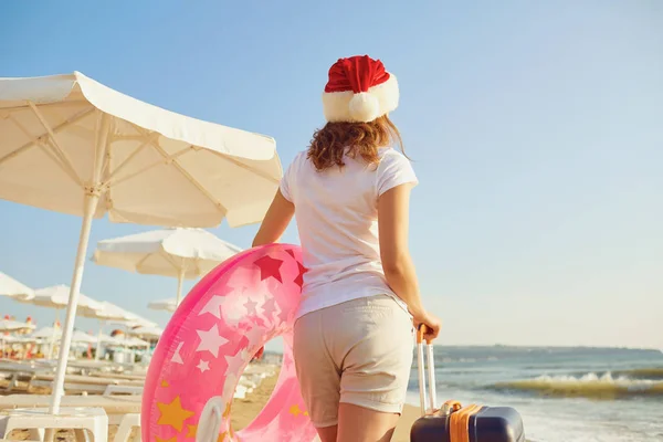 Dziewczyna w kapeluszu Mikołaja z walizką na plaży na Chri — Zdjęcie stockowe