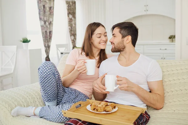 Um jovem casal com canecas e comida em uma bandeja sentar-se na sala . — Fotografia de Stock