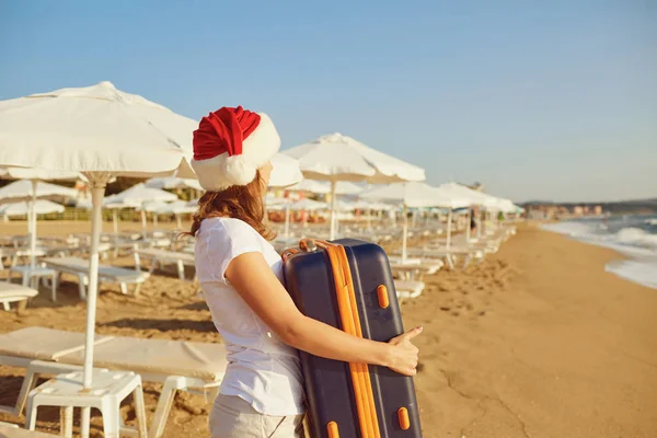 Dziewczyna w kapeluszu Mikołaja z walizką na plaży na Chri — Zdjęcie stockowe