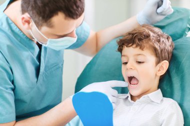 Erkek çocuk ve dişçi aynaya diş kontrol ediyor.