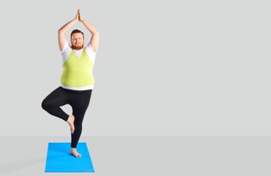 Komik şişman adam gri bir arka plan üzerinde duran yoga egzersizleri yapıyor.