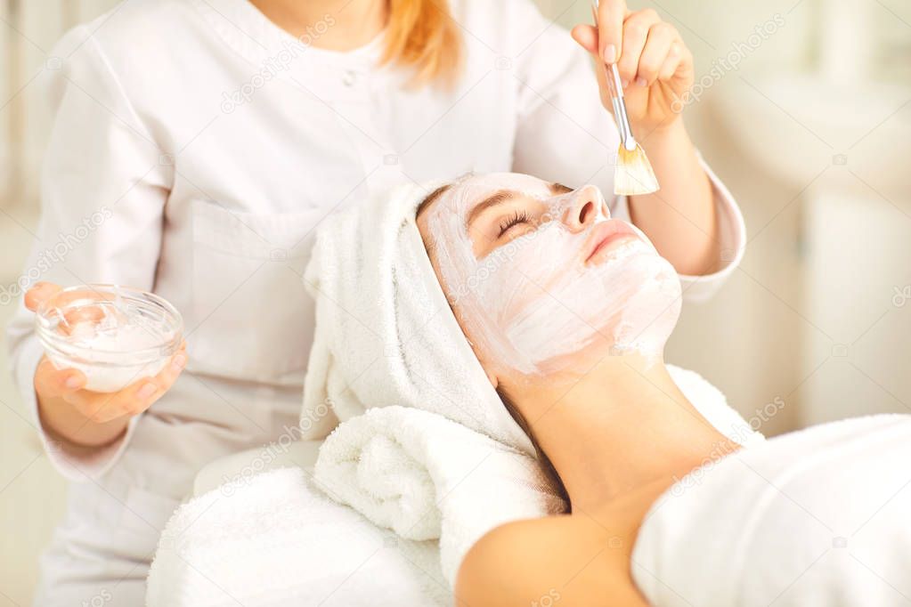 Beautician applies a white mask to a woman in the spa salon.