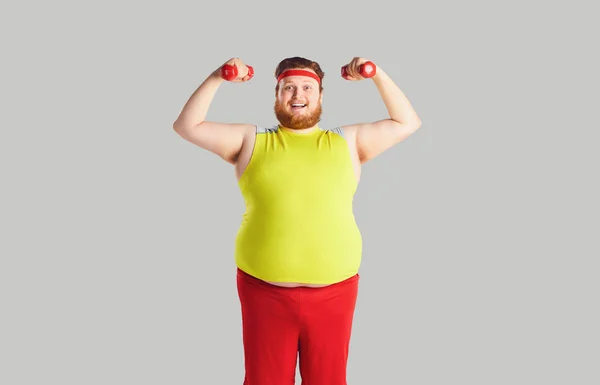 De dikke grappige man met een baard met dumbbells op een grijze achtergrond. — Stockfoto