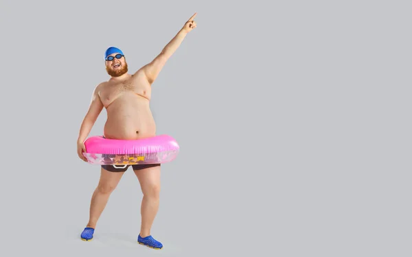 Fat funny man in swimming trunks with an inflatable ring on a gray background.