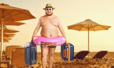 Bir yazlık plajın arka planına karşı bir bavul ve lastik yüzük ile mayo kalın adam.