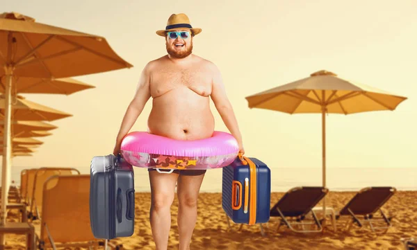 Dikke man in een badpak met een koffer en een rubberen ring tegen de achtergrond van een zomer strand. — Stockfoto