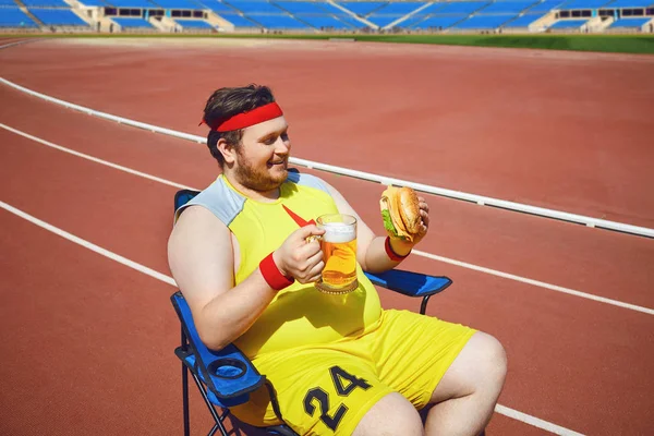 Un barbat gras mananca un burger si bere la antrenament pe stadion . — Fotografie, imagine de stoc