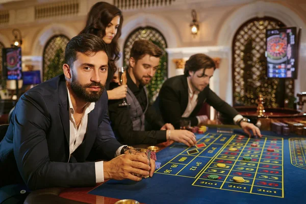 Un hombre con un traje con un vaso de whisky sentado en la ruleta de la mesa jugando al póquer en un casino . — Foto de Stock