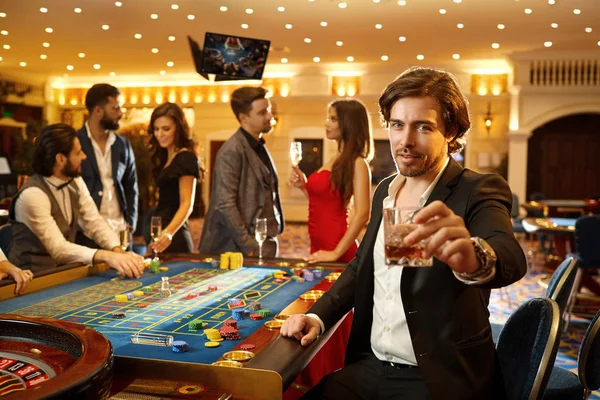 Un hombre con un traje sostiene una copa con alcohol en la mano contra el fondo de un juego de ruleta en un casino . —  Fotos de Stock