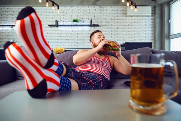 ソファに座ってハンバーガーを食べて厚い変な男. — ストック写真