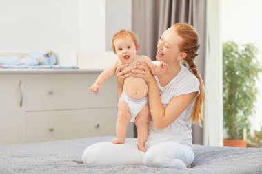 Mutlu anne odada yatakta oturan bebek gülümsüyor.