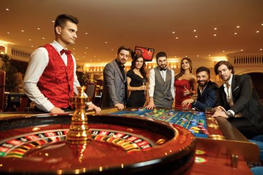 Group happy friends make bets gambiling at the roulette table in the casino. clipart