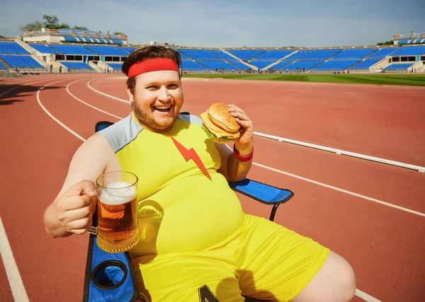 Un barbat gras mananca un burger si bere la antrenament pe stadion . — Fotografie, imagine de stoc