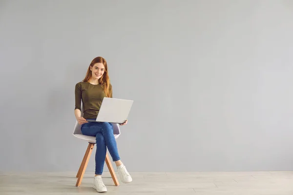 Рыжеволосая красивая студентка с ноутбуком сидит на стуле на сером фоне . — стоковое фото