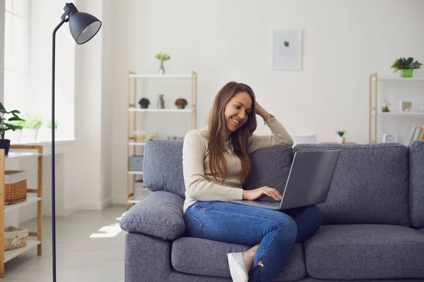 Online chat. Young woman works with a laptop using a web video camera while sitting on a sofa in a room. — Stock Photo, Image