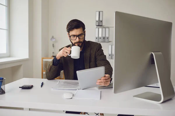 Trabalhador de negócios bebendo uma caneca de café enquanto sentado em uma mesa com um computador trabalhando no escritório . — Fotografia de Stock