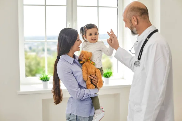 Családi orvos. A rangidős orvos pacsit ad a lánynak az orvosi rendelőben. Egy kis türelem az anyukájával a gyermekorvosnál. — Stock Fotó