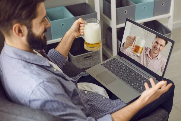 Un uomo barbuto con un bicchiere di birra si sta divertendo a parlare con un amico usando una videochiamata portatile a casa. Amici bere birra in remoto chat e avere una webcam chat . — Foto Stock