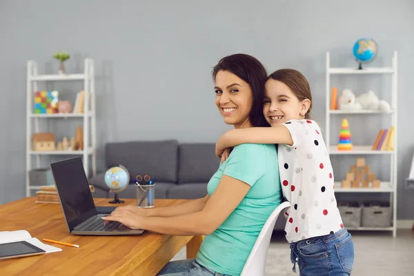 Прекрасна дівчина обіймає маму, працюючи онлайн через ноутбук вдома. Мила дитина обіймає свою матір в приміщенні — стокове фото