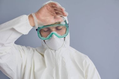 Gri arka planda tehlikeli madde kıyafeti giyen yorgun bir doktor. Bulaşıcı hastalıklardan bitap düşmüş bir tıp uzmanı.