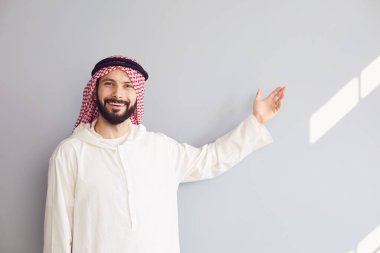 Çekici gülümseyen Arap adam elini gri bir arka plana doğrultuyor.