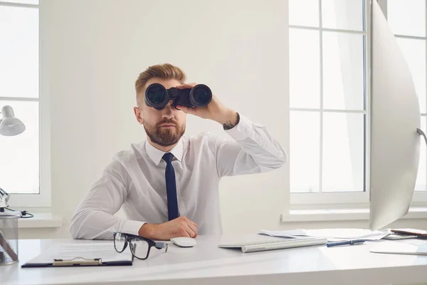 A businessman looks through binoculars while sitting at a table with a computer in the office. — ストック写真