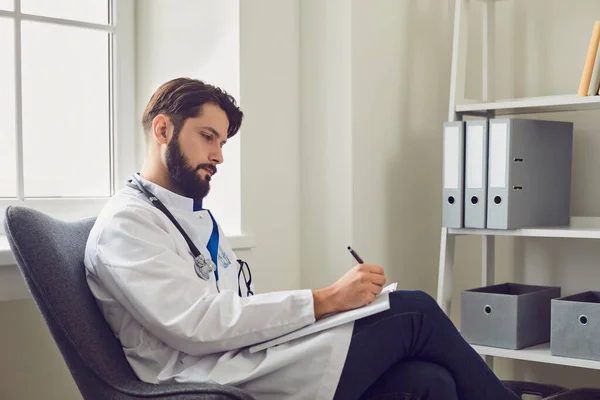 Seitenansicht junger männlicher Arzt schreibt Fallbericht in komfortabler Arztpraxis. — Stockfoto