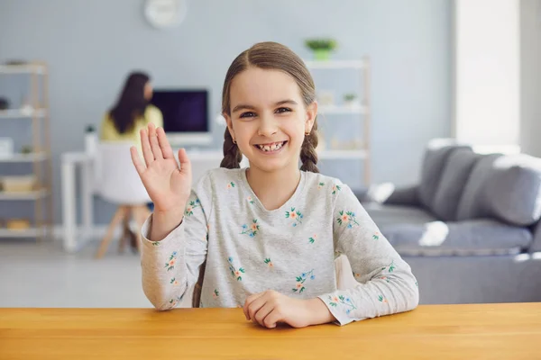 Дівчинка дивиться на веб-камеру, махаючи рукою у вітальні — стокове фото
