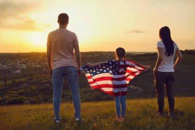 Mutlu aile, Amerika bayrağını gün batımında doğada taşıyor. Arkadan bak. Anne baba ve kızı Amerika 'nın Bağımsızlık Günü' nü kutluyor..