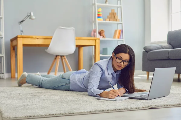 Arbeit zu Hause. Schöne Frau mit Brille macht sich Notizen während einer Online-Business-Konferenz oder eines Bildungskurses auf dem Laptop im Zimmer — Stockfoto