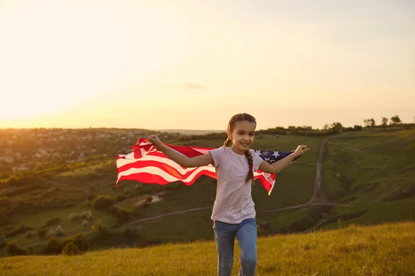 Gadis dengan bendera Amerika berjalan melalui lapangan saat matahari terbenam di alam. — Stok Foto