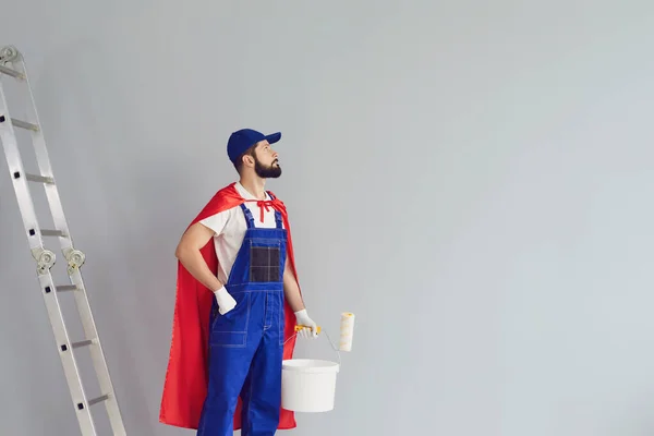 快乐的家庭修理工在灰色背景下从事房屋翻新工作.年轻的承包商拿着滚筒刷，穿着超级英雄的服装接近灰色墙壁. — 图库照片