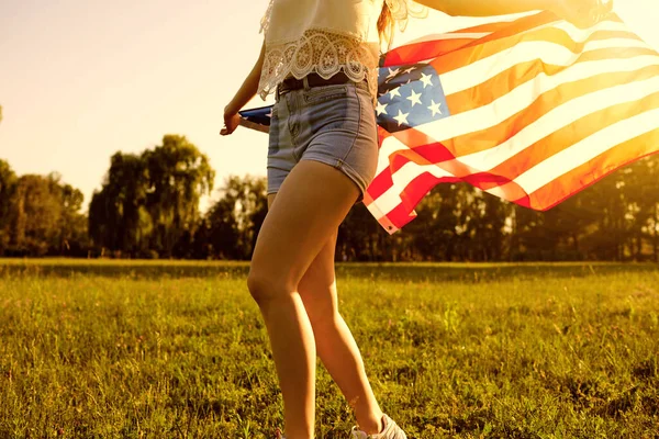 Independence Day USA. Girl in shorts with an American flag runs on the grass in a summer park at sunset.