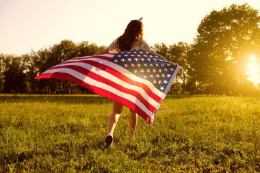 Arkadan bak. Amerikan bayrağı taşıyan bir kız gün batımında doğada koşar. ABD Bağımsızlık Günü.